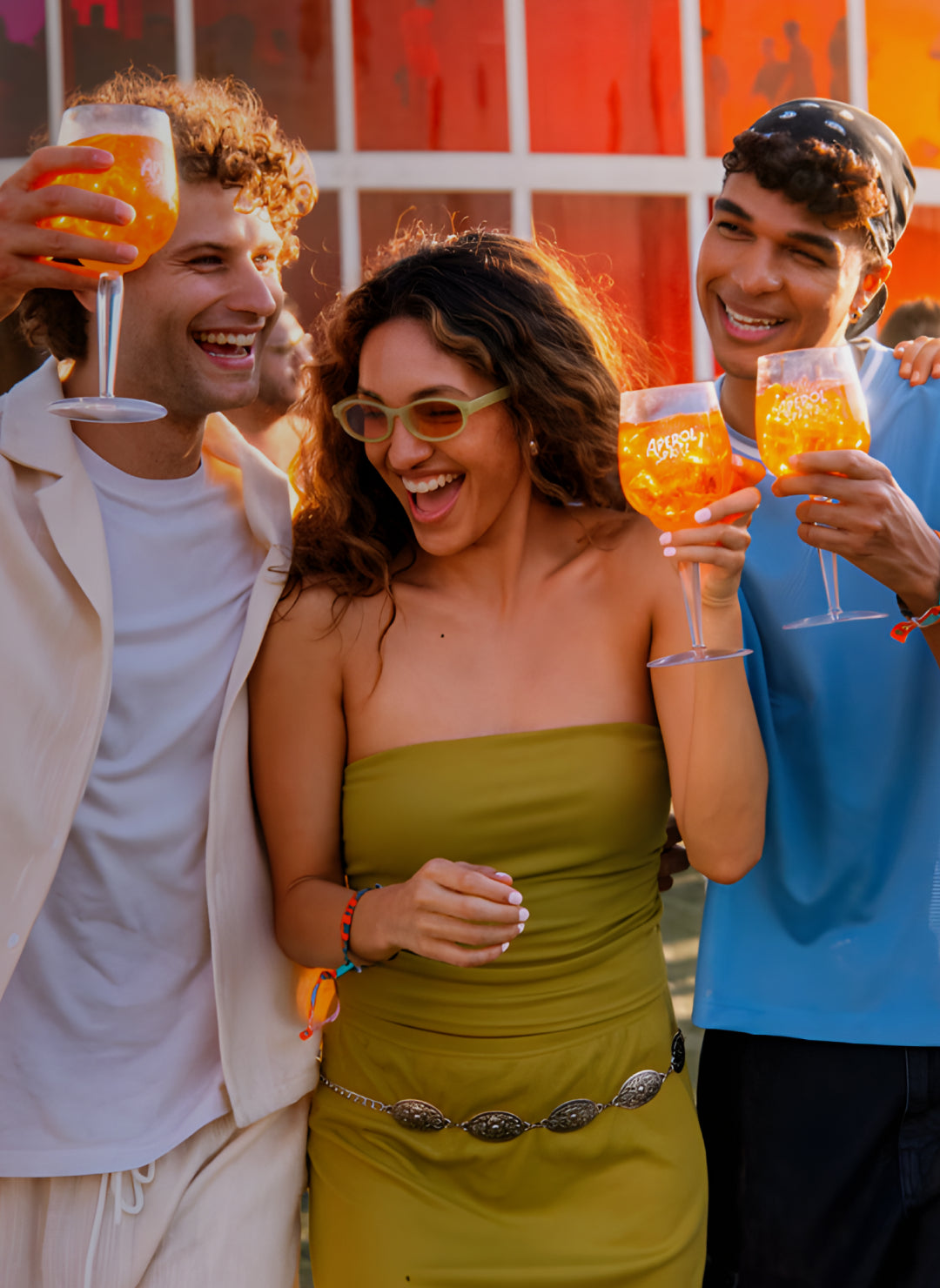 Three friends smiling and holding Aperol Spritz at an outdoor festival, dressed in summer outfits